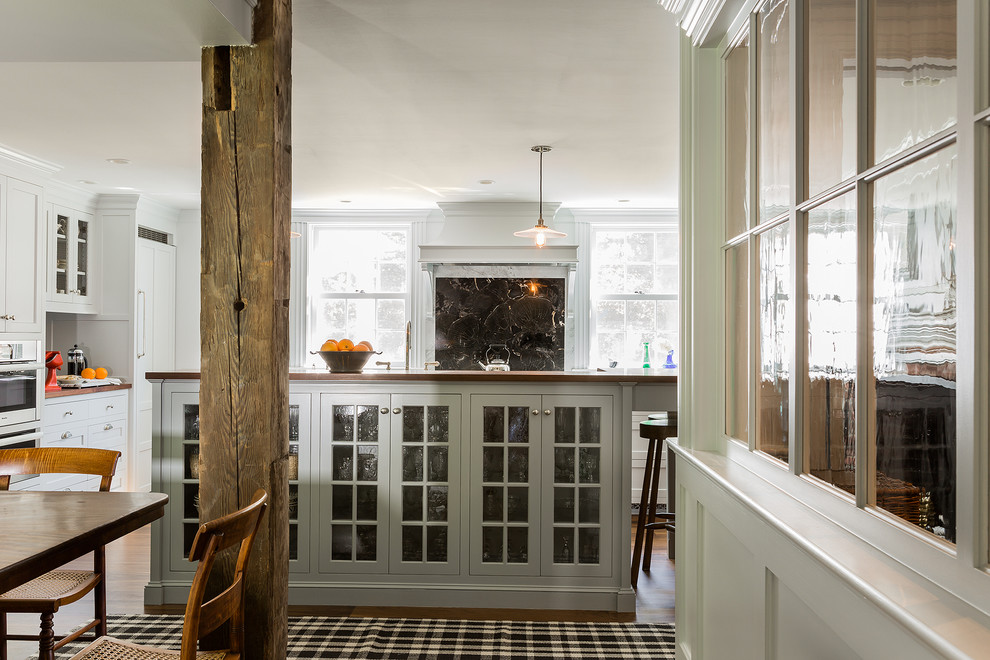 Country eat-in kitchen in Boston with a farmhouse sink, glass-front cabinets, grey cabinets, wood benchtops, black splashback, stone slab splashback, panelled appliances, medium hardwood floors and with island.