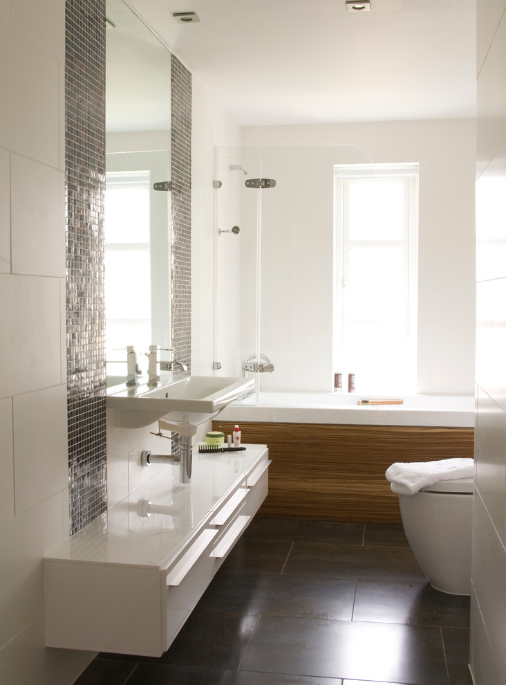 Photo of a mid-sized contemporary master bathroom in Sussex with white cabinets, a drop-in tub, a shower/bathtub combo, porcelain tile, white walls, porcelain floors, a wall-mount sink, flat-panel cabinets, a one-piece toilet, white tile and brown floor.