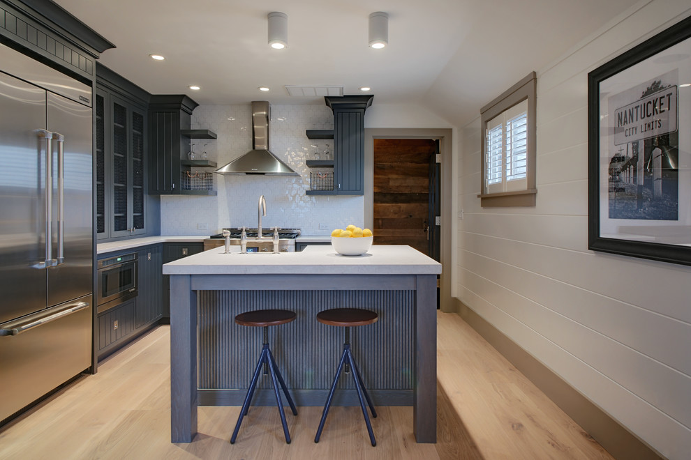 Photo of a beach style l-shaped separate kitchen in Boston with quartz benchtops, porcelain splashback, glass-front cabinets, blue cabinets, white splashback, stainless steel appliances, light hardwood floors, with island and beige floor.