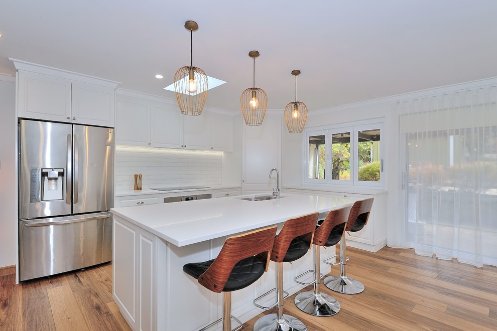 Mid-sized contemporary l-shaped open plan kitchen in Perth with a double-bowl sink, shaker cabinets, white cabinets, quartz benchtops, white splashback, subway tile splashback, stainless steel appliances, laminate floors, with island, brown floor and white benchtop.