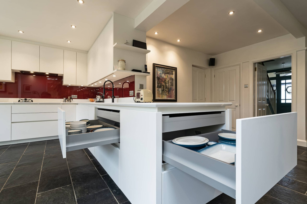 This is an example of a mid-sized traditional l-shaped eat-in kitchen in Oxfordshire with an integrated sink, white cabinets, red splashback, glass sheet splashback, stainless steel appliances, ceramic floors, no island, black floor and white benchtop.