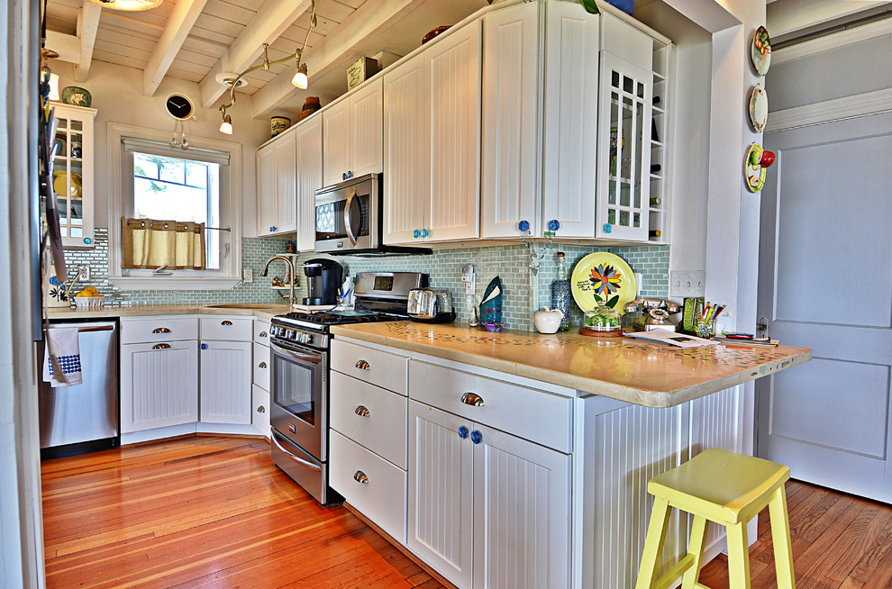 This is an example of a mid-sized beach style l-shaped kitchen in DC Metro with a drop-in sink, beaded inset cabinets, white cabinets, concrete benchtops, blue splashback, glass tile splashback, stainless steel appliances and medium hardwood floors.