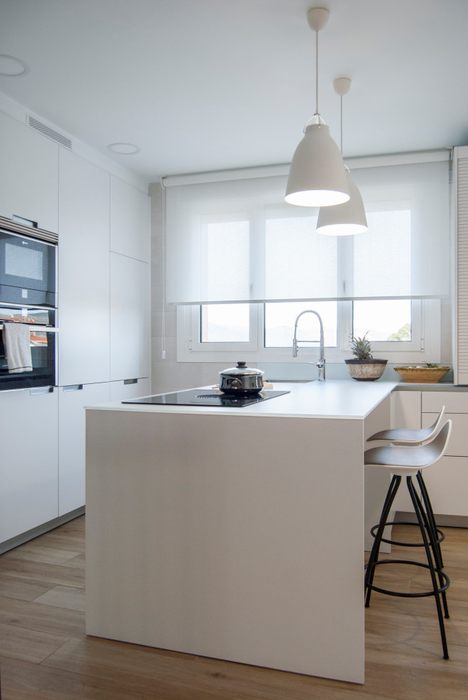 Photo of a mid-sized contemporary l-shaped open plan kitchen in Bilbao with an undermount sink, flat-panel cabinets, white cabinets, glass benchtops, white splashback, black appliances, medium hardwood floors, a peninsula and white benchtop.