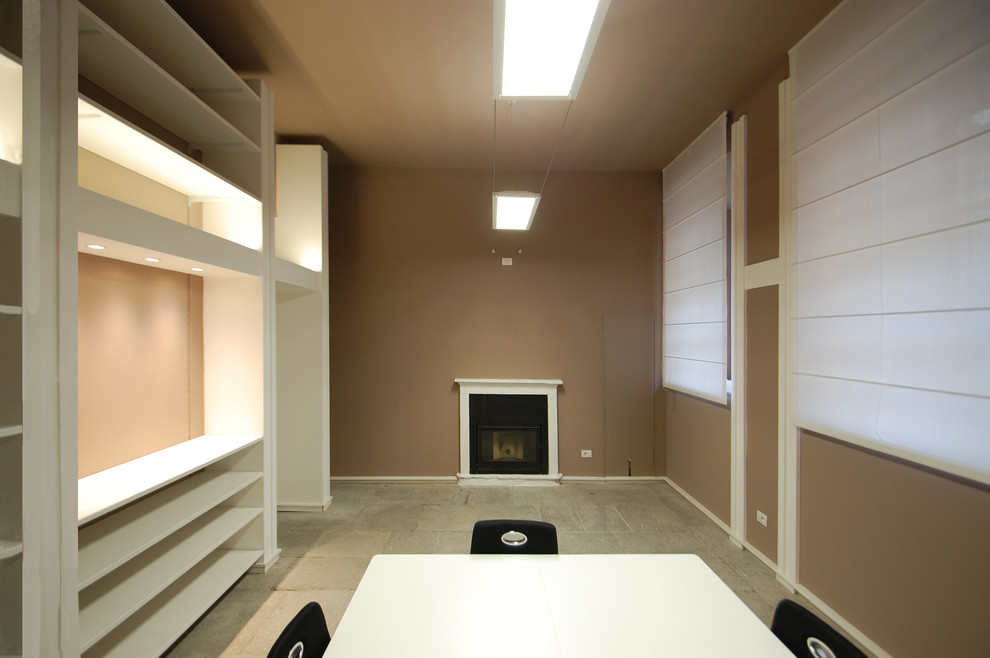 This is an example of a large contemporary home office in Milan with a library, brown walls, limestone floors, a standard fireplace, a concrete fireplace surround, a freestanding desk and grey floor.