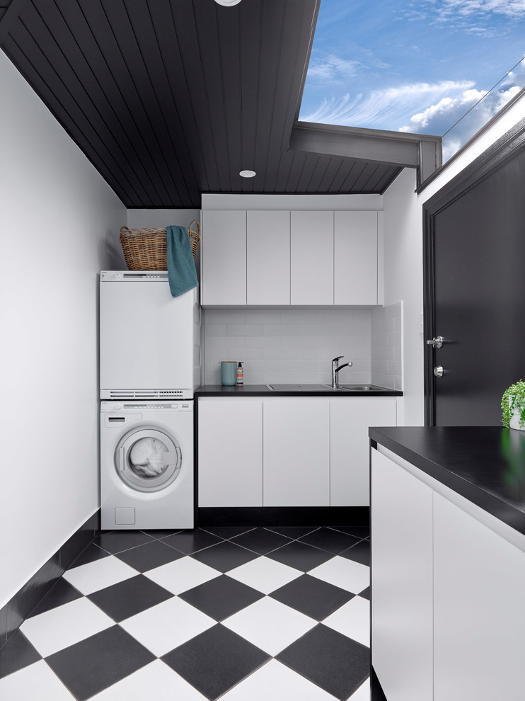 Mid-sized transitional l-shaped dedicated laundry room with a drop-in sink, flat-panel cabinets, white cabinets, laminate benchtops, white walls, porcelain floors, a stacked washer and dryer, multi-coloured floor and black benchtop.