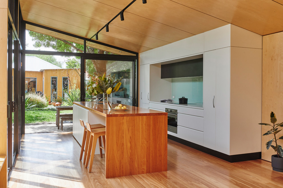 Photo of a contemporary galley kitchen in Melbourne with a double-bowl sink, flat-panel cabinets, white cabinets, wood benchtops, blue splashback, glass sheet splashback, panelled appliances, medium hardwood floors, with island and brown floor.