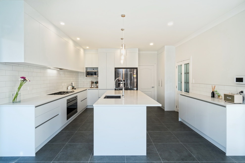 Modern l-shaped separate kitchen in Auckland with an undermount sink, flat-panel cabinets, white cabinets, white splashback, subway tile splashback, stainless steel appliances, with island, black floor and white benchtop.