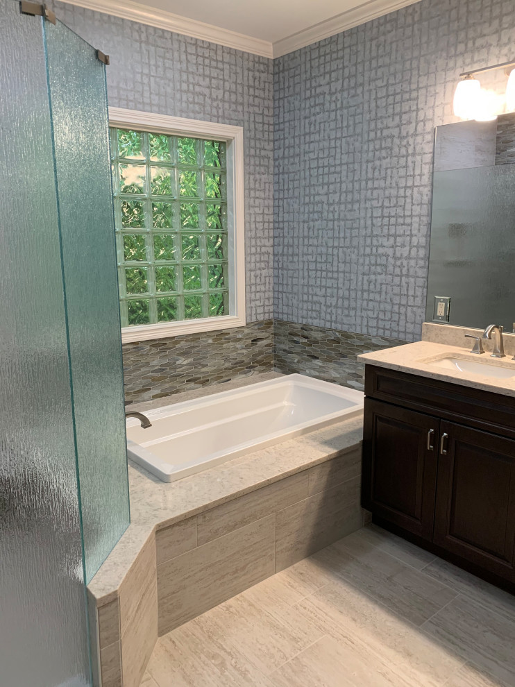 Photo of a mid-sized transitional master wet room bathroom in Cincinnati with raised-panel cabinets, dark wood cabinets, a drop-in tub, a two-piece toilet, multi-coloured tile, glass tile, multi-coloured walls, an undermount sink, engineered quartz benchtops, beige floor, beige benchtops, a niche, a double vanity, a built-in vanity and wallpaper.
