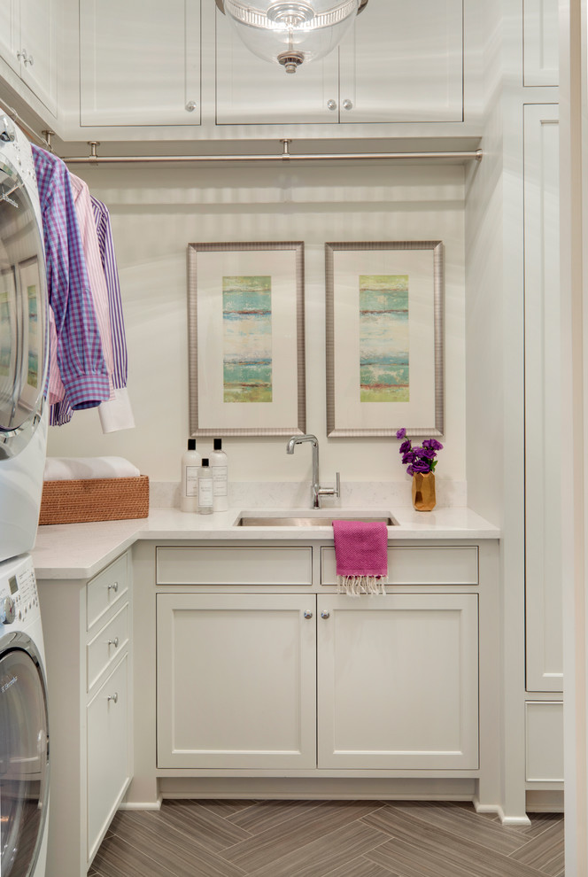Traditional l-shaped laundry room in Minneapolis with an undermount sink, white cabinets, solid surface benchtops, white walls, ceramic floors, a stacked washer and dryer and shaker cabinets.
