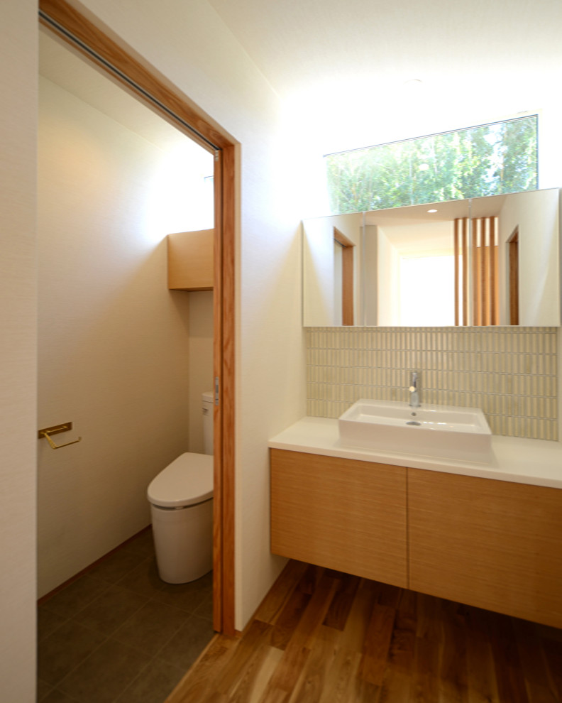 Foto på ett mellanstort vit toalett, med vita väggar, mellanmörkt trägolv, brunt golv, släta luckor, vita skåp, en toalettstol med separat cisternkåpa, beige kakel, mosaik, ett fristående handfat och bänkskiva i akrylsten