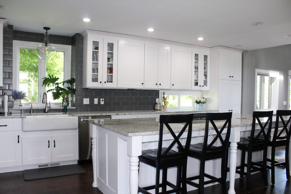 Large transitional l-shaped brown floor eat-in kitchen photo with a farmhouse sink, shaker cabinets, white cabinets, quartz countertops, gray backsplash, subway tile backsplash, stainless steel appliances, an island and multicolored countertops