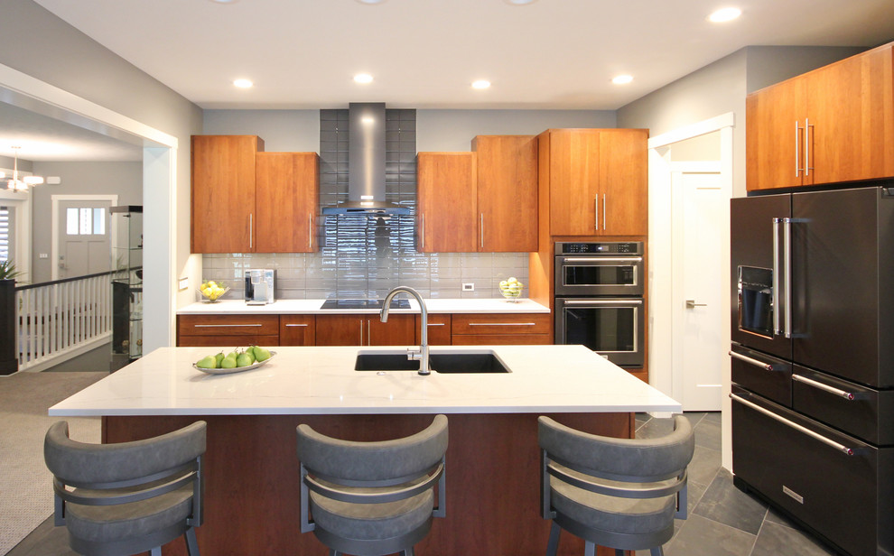 Photo of a mid-sized contemporary l-shaped eat-in kitchen in Grand Rapids with an undermount sink, flat-panel cabinets, brown cabinets, quartz benchtops, grey splashback, subway tile splashback, black appliances, ceramic floors and with island.