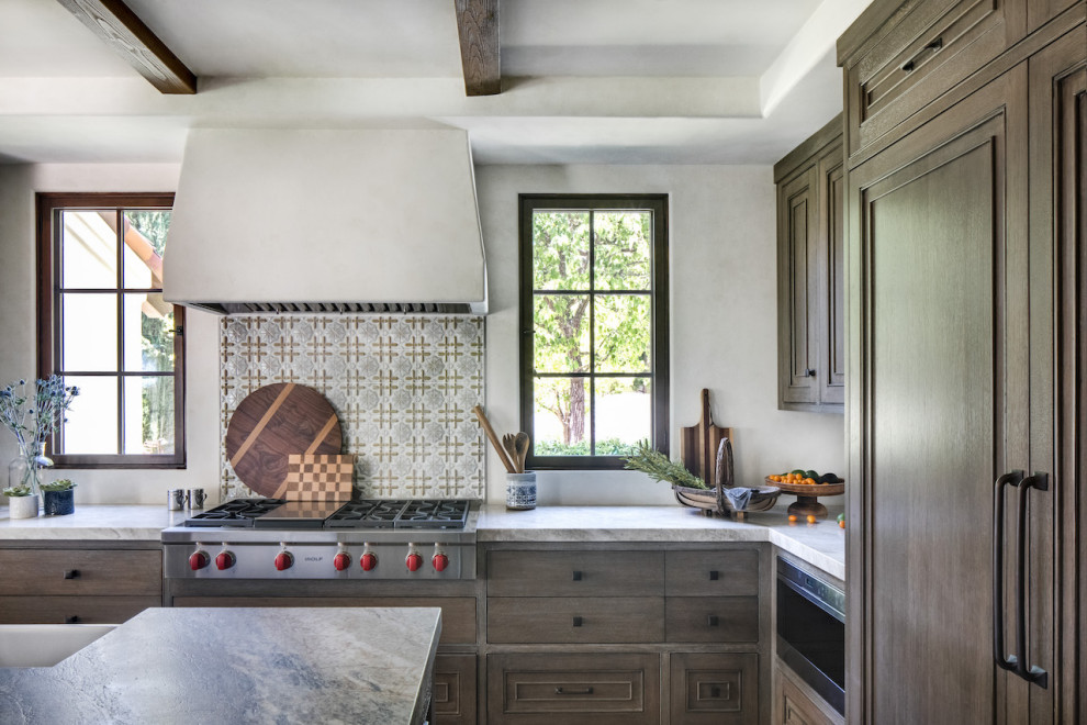 Photo of a large mediterranean u-shaped eat-in kitchen in Orange County with a farmhouse sink, raised-panel cabinets, medium wood cabinets, limestone benchtops, blue splashback, ceramic splashback, panelled appliances, travertine floors, with island, beige floor and grey benchtop.