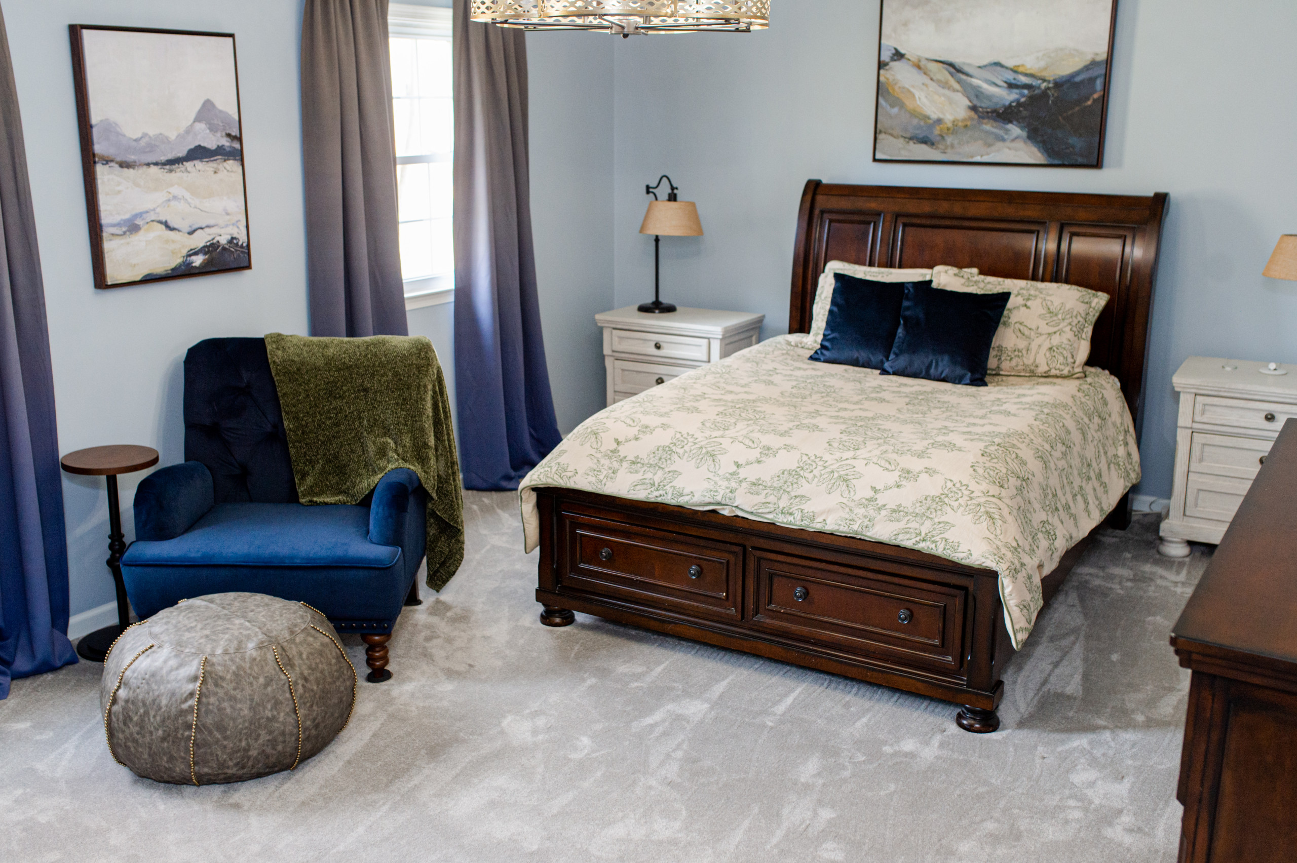 Traditional style Owner's Bedroom with queen size bed, seating area with pouf ottoman, accent color nightstands, fandelier, and navy blue and olive green accent colors