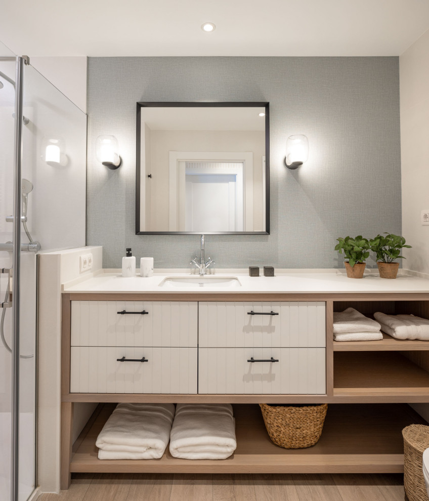 This is an example of a large beach style master bathroom in Bilbao with white cabinets, a curbless shower, a wall-mount toilet, white tile, porcelain tile, blue walls, laminate floors, an undermount sink, engineered quartz benchtops, a hinged shower door, beige benchtops, a single vanity, a built-in vanity, wallpaper and flat-panel cabinets.