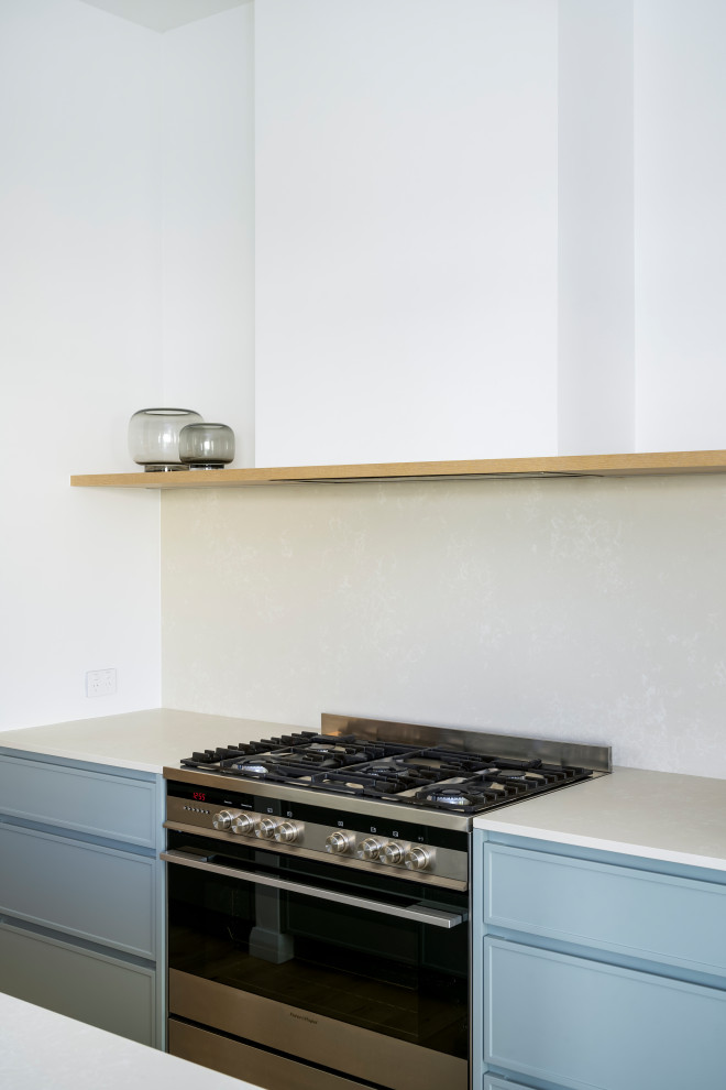 Modern inredning av ett mellanstort vit vitt kök, med en undermonterad diskho, skåp i shakerstil, blå skåp, bänkskiva i kvarts, vitt stänkskydd, rostfria vitvaror, ljust trägolv och en halv köksö
