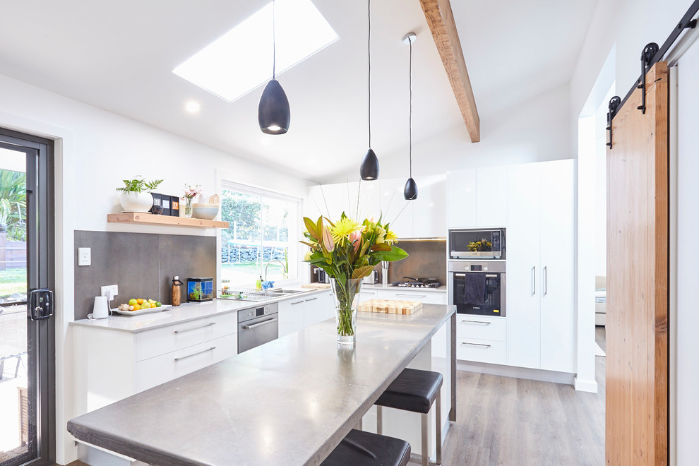 Inspiration for a contemporary l-shaped kitchen in Central Coast with a double-bowl sink, flat-panel cabinets, white cabinets, stainless steel benchtops, grey splashback, black appliances, light hardwood floors, with island, brown floor and grey benchtop.