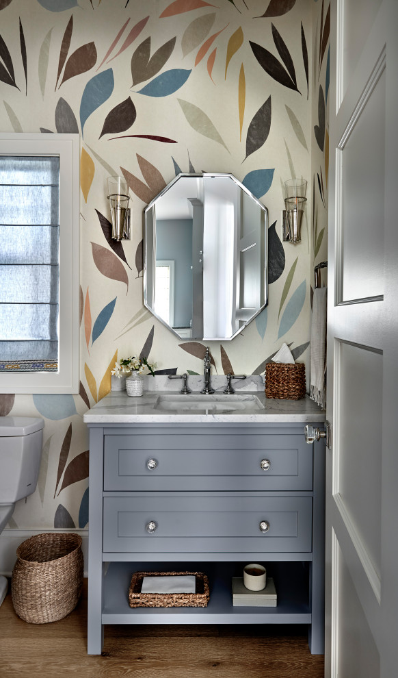 Design ideas for a mid-sized beach style powder room in Chicago with recessed-panel cabinets, blue cabinets, light hardwood floors, a drop-in sink, marble benchtops, brown floor, white benchtops, a freestanding vanity, wallpaper, a two-piece toilet and multi-coloured walls.