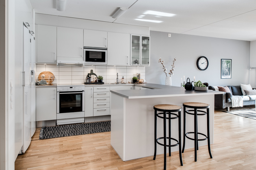 Mid-sized scandinavian l-shaped open plan kitchen in Gothenburg with flat-panel cabinets, white cabinets, white splashback, white appliances, light hardwood floors, a peninsula and a single-bowl sink.