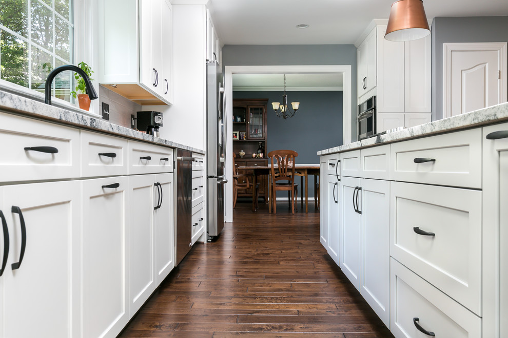 This is an example of a mid-sized transitional galley open plan kitchen in DC Metro with an undermount sink, shaker cabinets, white cabinets, granite benchtops, grey splashback, porcelain splashback, stainless steel appliances, with island, brown floor and dark hardwood floors.