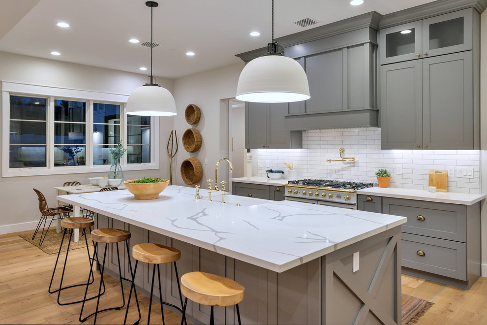 Photo of a large arts and crafts single-wall open plan kitchen in Phoenix with a farmhouse sink, shaker cabinets, grey cabinets, quartzite benchtops, white splashback, subway tile splashback, stainless steel appliances, light hardwood floors, with island, brown floor and white benchtop.