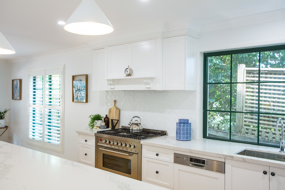 This is an example of an expansive traditional galley eat-in kitchen in Brisbane with a single-bowl sink, shaker cabinets, white cabinets, solid surface benchtops, white splashback, subway tile splashback, stainless steel appliances, medium hardwood floors, with island, brown floor and white benchtop.