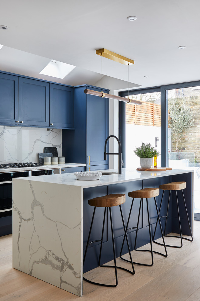 Inspiration for a transitional kitchen in London with an undermount sink, shaker cabinets, blue cabinets, white splashback, black appliances, light hardwood floors, with island, beige floor and white benchtop.