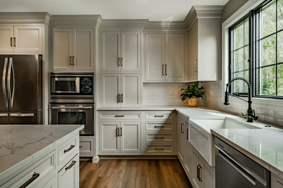 Example of a mid-sized classic l-shaped brown floor eat-in kitchen design in Philadelphia with a farmhouse sink, flat-panel cabinets, beige cabinets, quartz countertops, white backsplash, ceramic backsplash, stainless steel appliances, an island and white countertops