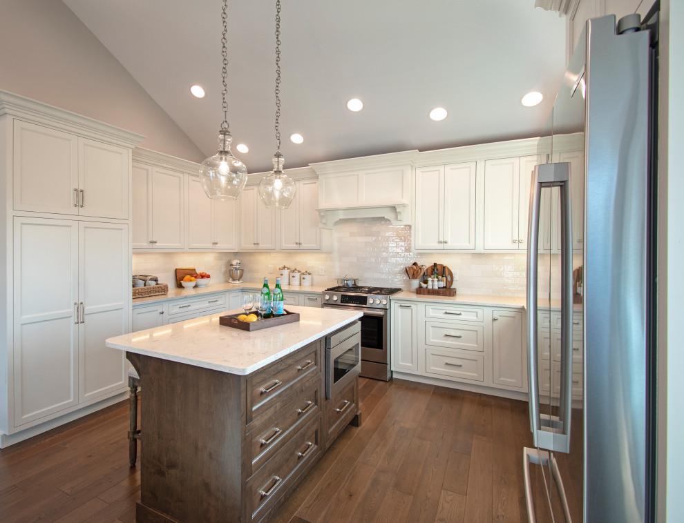Example of a transitional medium tone wood floor, brown floor and vaulted ceiling kitchen pantry design with an undermount sink, shaker cabinets, white cabinets, quartz countertops, white backsplash, ceramic backsplash, stainless steel appliances, an island and white countertops