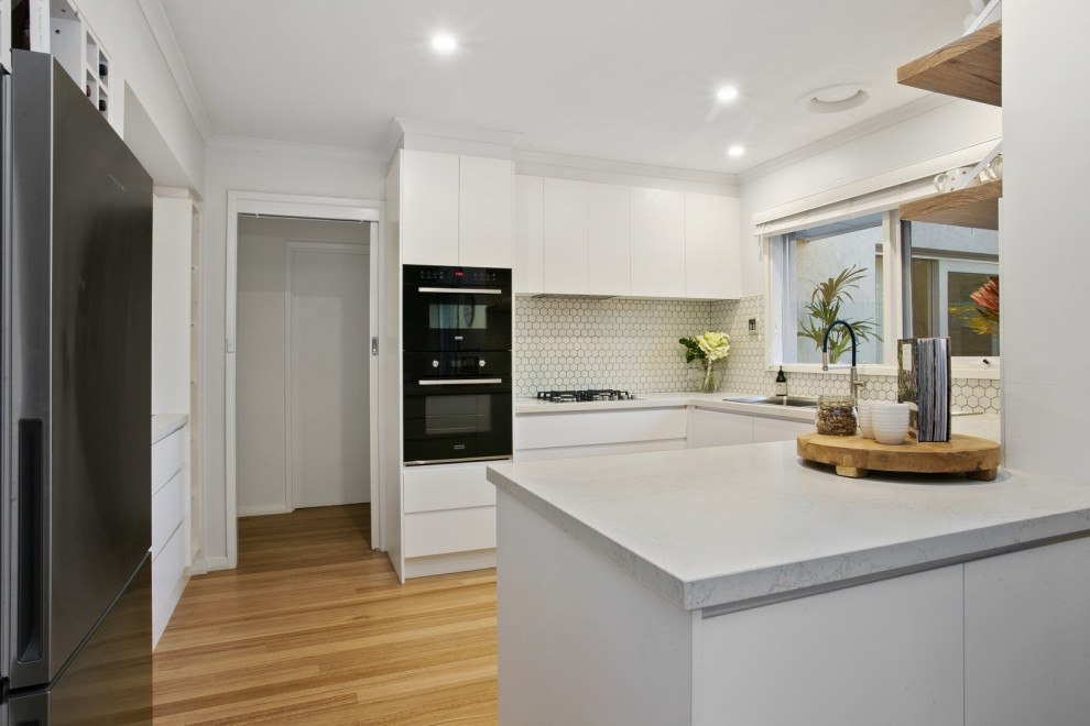 This is an example of a mid-sized beach style u-shaped eat-in kitchen in Geelong with a double-bowl sink, white cabinets, soapstone benchtops, white splashback, ceramic splashback, stainless steel appliances, light hardwood floors and with island.