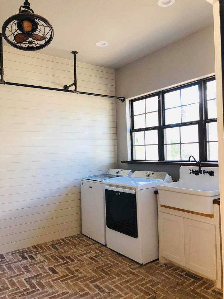 Inspiration for a mid-sized country single-wall utility room in Austin with a farmhouse sink, shaker cabinets, white cabinets, white walls, brick floors, a side-by-side washer and dryer and brown floor.
