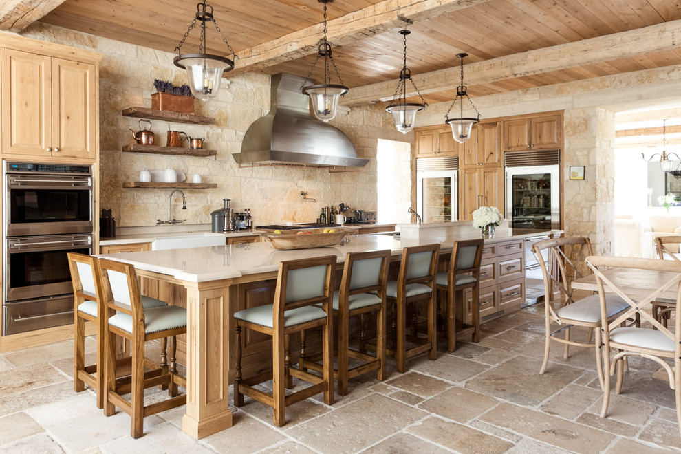 Photo of a mediterranean l-shaped eat-in kitchen in Dallas with a farmhouse sink, raised-panel cabinets, medium wood cabinets, beige splashback, stone tile splashback, stainless steel appliances and with island.