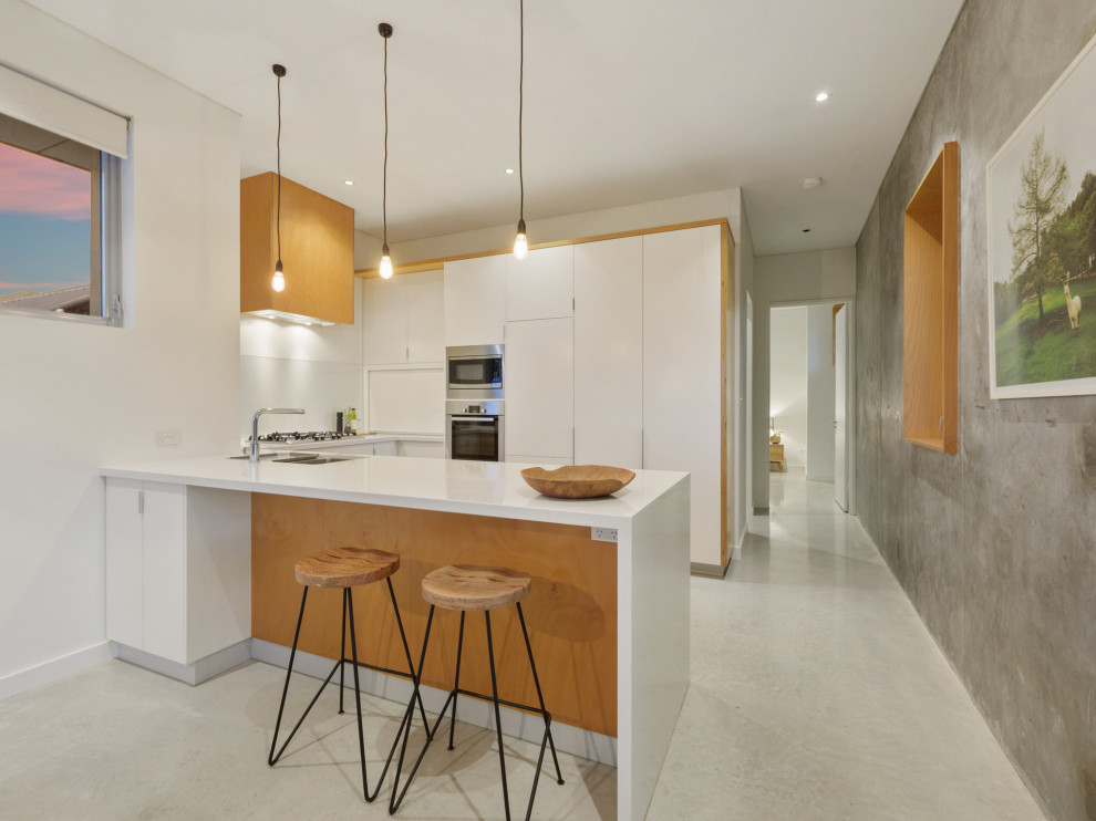 Photo of a midcentury u-shaped kitchen in Perth with a double-bowl sink, flat-panel cabinets, white cabinets, stainless steel appliances, concrete floors, a peninsula, grey floor and white benchtop.