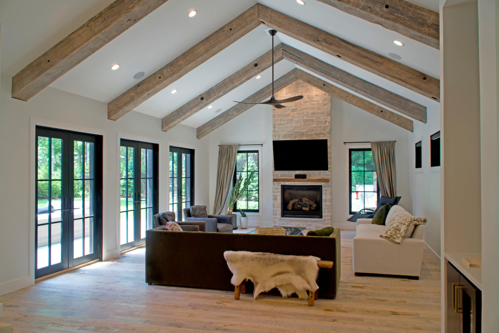 Example of a cottage open concept medium tone wood floor, beige floor and exposed beam family room design with white walls, a standard fireplace, a stone fireplace and a wall-mounted tv