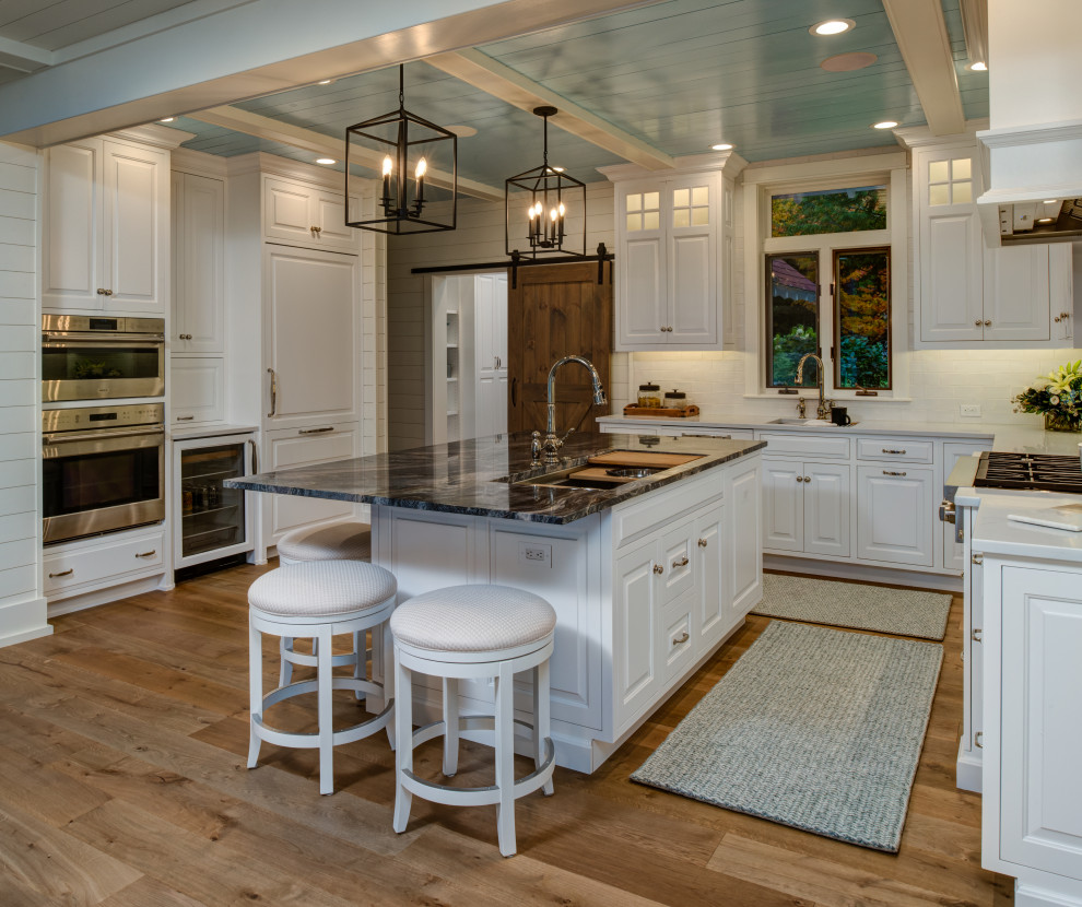 This is an example of a large beach style l-shaped eat-in kitchen in Other with recessed-panel cabinets, white cabinets, quartz benchtops, white splashback, ceramic splashback, panelled appliances, medium hardwood floors, with island, brown floor and white benchtop.