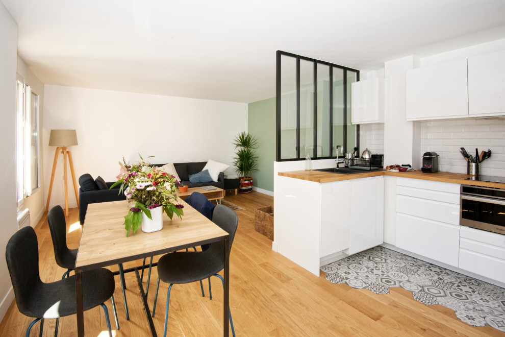 Design ideas for a mid-sized scandinavian l-shaped eat-in kitchen in Bordeaux with a drop-in sink, flat-panel cabinets, white cabinets, wood benchtops, white splashback, subway tile splashback, black appliances, light hardwood floors, no island, beige floor and beige benchtop.