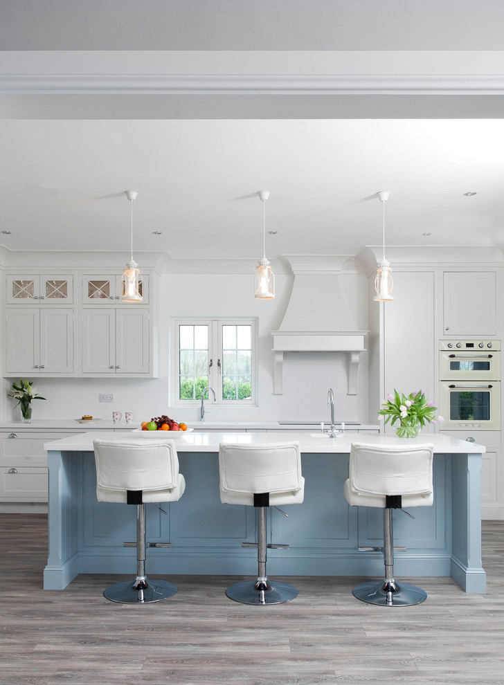 Photo of a large beach style kitchen in Other with white cabinets, quartzite benchtops, white splashback, light hardwood floors, with island, white benchtop, white appliances and shaker cabinets.