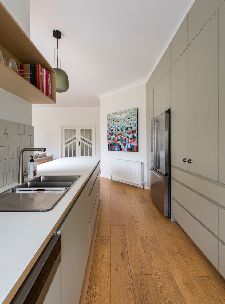 This is an example of a contemporary u-shaped kitchen/diner in Melbourne with a double-bowl sink, flat-panel cabinets, beige cabinets, composite countertops, beige splashback, ceramic splashback, stainless steel appliances, medium hardwood flooring, an island, brown floors and white worktops.