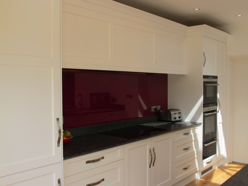 Medium sized contemporary l-shaped open plan kitchen in Surrey with a built-in sink, flat-panel cabinets, granite worktops, red splashback, glass tiled splashback, stainless steel appliances, medium hardwood flooring and an island.