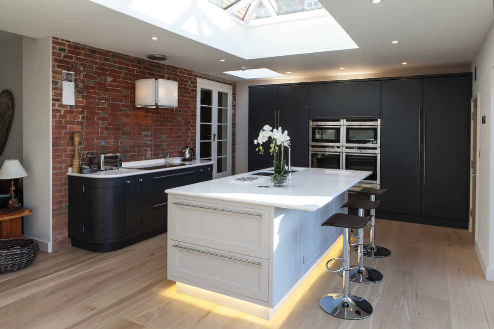 Design ideas for a contemporary l-shaped kitchen in Hampshire with a double-bowl sink, flat-panel cabinets, grey cabinets, glass sheet splashback, panelled appliances, light hardwood floors and with island.