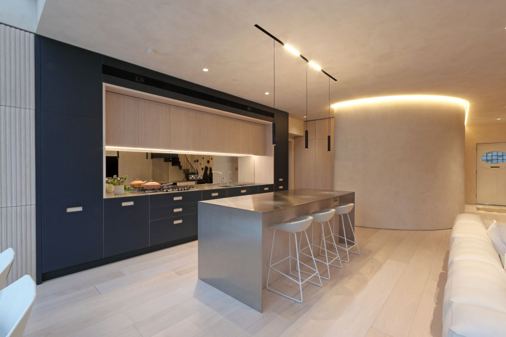 This is an example of a large modern galley kitchen/diner in London with an integrated sink, blue cabinets, stainless steel worktops, mirror splashback, integrated appliances, light hardwood flooring, an island, beige floors and feature lighting.