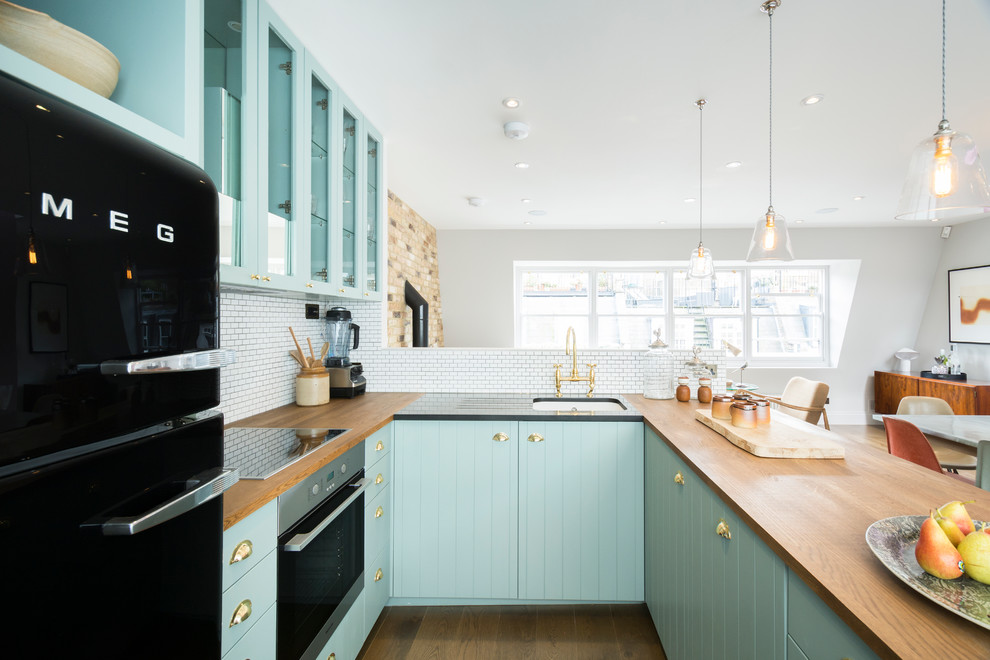 Small country u-shaped open plan kitchen in London with glass-front cabinets, blue cabinets, wood benchtops, a peninsula, an undermount sink, white splashback, black appliances and dark hardwood floors.