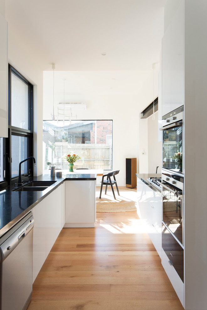 Photo of a mid-sized arts and crafts galley eat-in kitchen in Melbourne with a drop-in sink, flat-panel cabinets, white cabinets, granite benchtops, black splashback, stone slab splashback, light hardwood floors and no island.