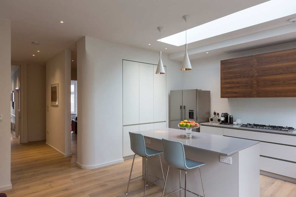 This is an example of a mid-sized contemporary l-shaped open plan kitchen in London with flat-panel cabinets, white cabinets, quartzite benchtops, white splashback, glass sheet splashback, light hardwood floors, with island, beige floor and white benchtop.