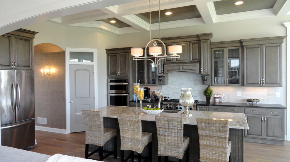 Example of a large transitional l-shaped light wood floor open concept kitchen design in Columbus with a farmhouse sink, raised-panel cabinets, gray cabinets, granite countertops, white backsplash, stone tile backsplash, stainless steel appliances and an island
