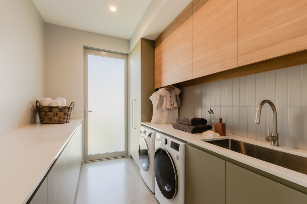 This is an example of a large contemporary u-shaped utility room in Sunshine Coast with an integrated sink, light wood cabinets, laminate benchtops, grey splashback, subway tile splashback, a side-by-side washer and dryer, white floor and white benchtop.