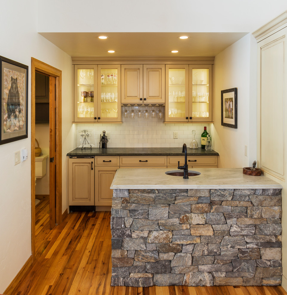 Design ideas for a mid-sized country wet bar in Denver with an undermount sink, beige cabinets, granite benchtops, white splashback, medium hardwood floors, glass-front cabinets, subway tile splashback and brown floor.