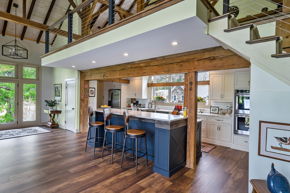 Example of a mid-sized cottage galley medium tone wood floor, brown floor and exposed beam open concept kitchen design in Philadelphia with a farmhouse sink, shaker cabinets, blue cabinets, quartz countertops, white backsplash, porcelain backsplash, stainless steel appliances, an island and white countertops