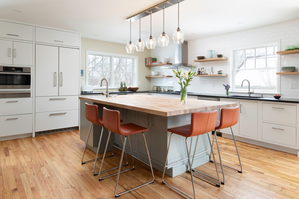 This is an example of a midcentury l-shaped kitchen in Denver with an undermount sink, flat-panel cabinets, white cabinets, white splashback, stainless steel appliances, medium hardwood floors, with island, brown floor and black benchtop.