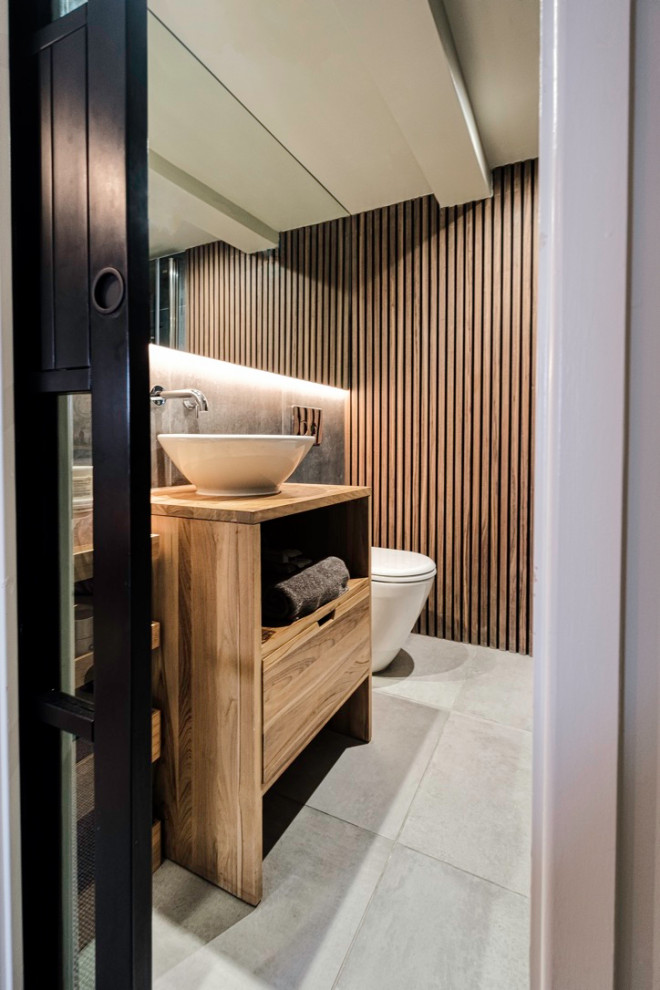 Photo of a small industrial ensuite bathroom in London with flat-panel cabinets, medium wood cabinets, a one-piece toilet, grey tiles, porcelain tiles, porcelain flooring, a vessel sink, wooden worktops, grey floors, a feature wall, a single sink, a freestanding vanity unit, exposed beams and panelled walls.